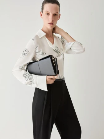 Model wearing a white floral blouse and black trousers holding a dark handbag against a seamless light background
