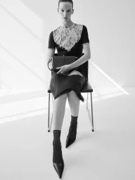 Model Sara Blomqvist seated on a chair wearing a black dress with white lace yoke, holding a handbag against a plain background