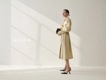 Model standing in profile wearing a beige belted trench coat and black heels against a light wall with diagonal shadows