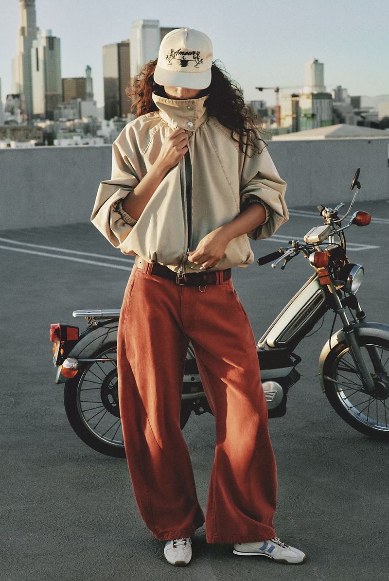 A person in a beige jacket and orange pants stands next to a moped on a rooftop parking lot with a city skyline.