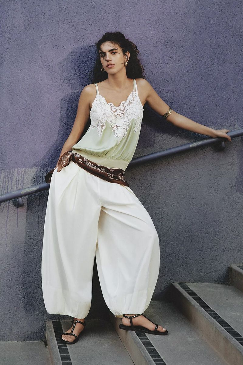 Woman in light green lace top and white palazzo pants poses on concrete stairs next to a metal handrail and purple wall.