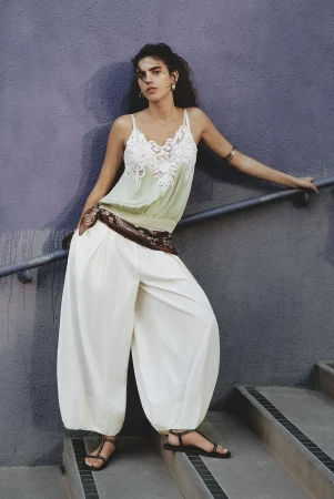 Model leaning against a handrail on outdoor steps wearing a light green camisole and wide white trousers.