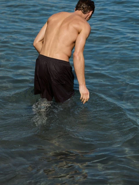 Back view of adult male standing in ocean water. He is wearing black shorts, and the water is up to his mid-thigh.