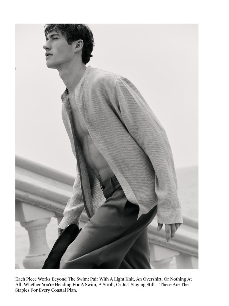 Monochrome fashion shot of a young adult male with fair skin, wearing a light knit shirt and pants, outdoors on a staircase.