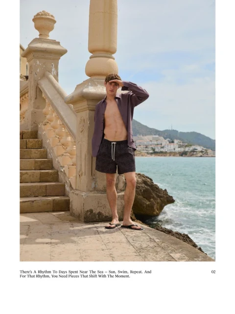 Male fashion model poses on a seaside walkway wearing patterned swim trunks and an open button-down shirt, against a coastal backdrop.