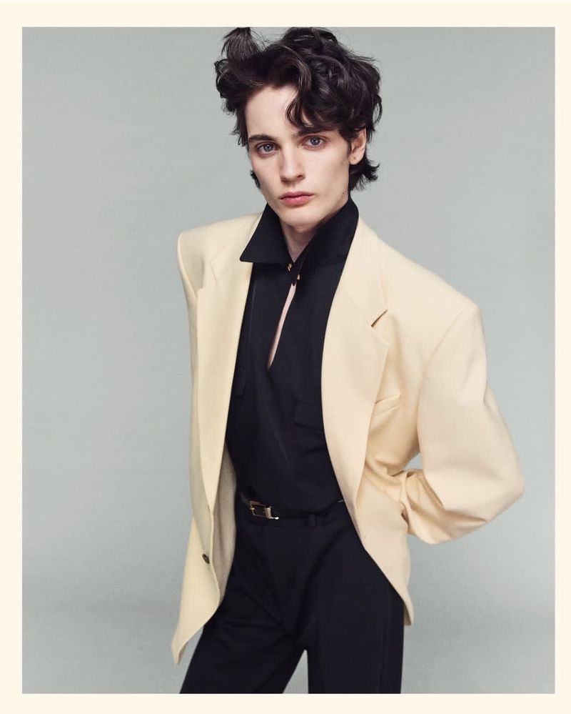 Adult male model in a studio shot, wearing a cream jacket with a black shirt and pants, showcasing a high-fashion aesthetic.