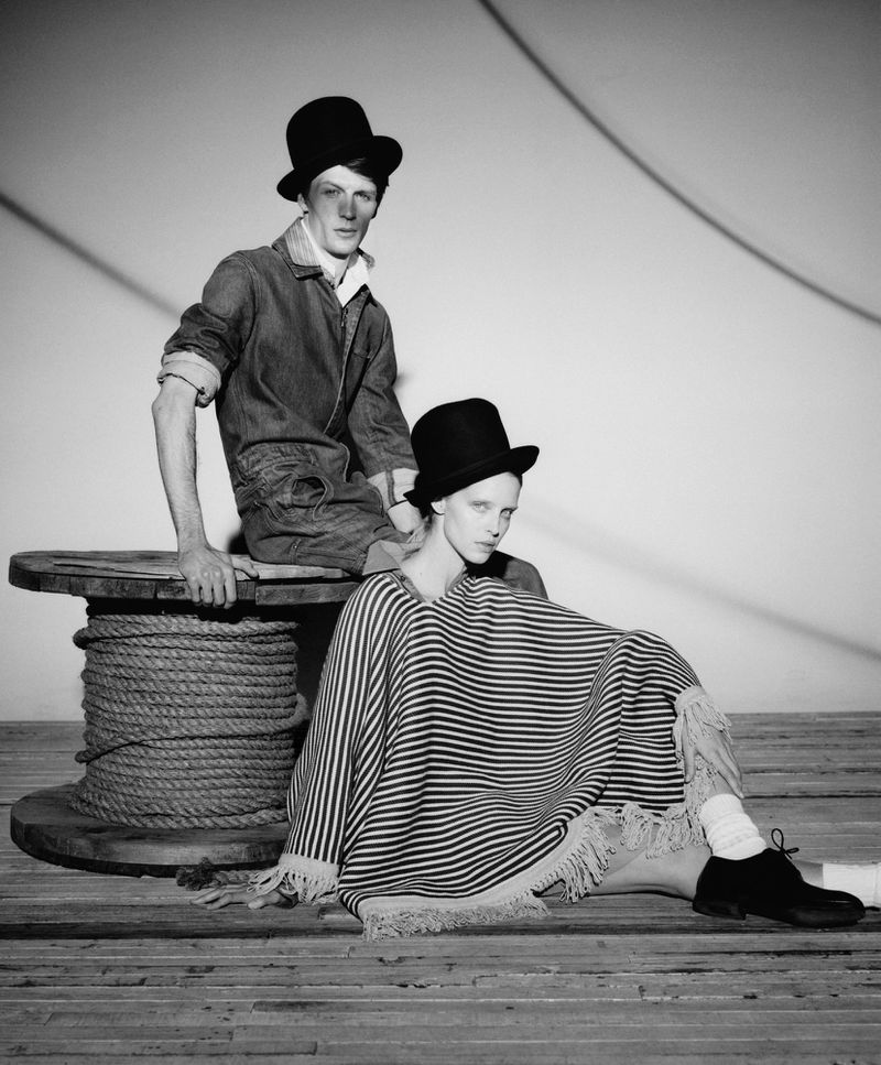 Black and white fashion shot featuring a man sitting on a wooden spool and a woman seated in front on a wood floor wearing a hat.