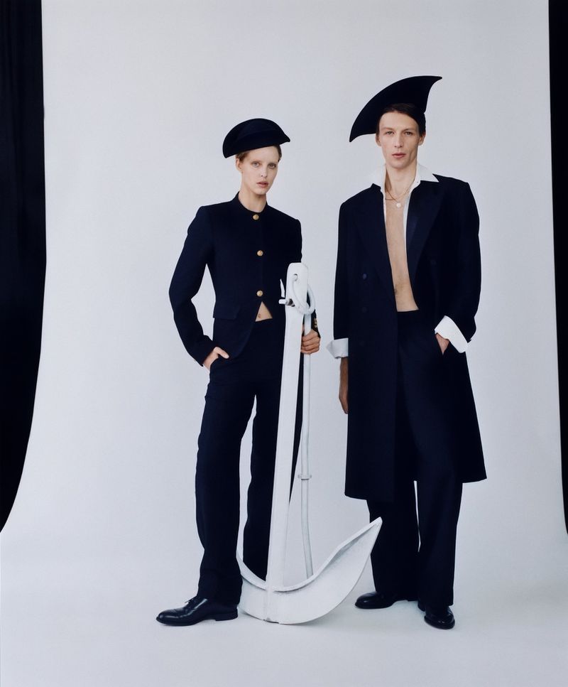 Two fashion models in matching navy suit ensembles and peaked hats pose indoors next to a large, white crescent-shaped prop.