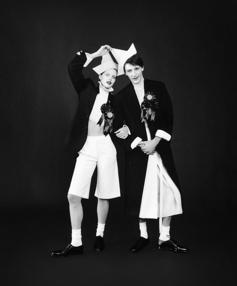 Two people in black and white, wearing matching outfits and paper hats, stand against a dark backdrop in a vintage-style fashion portrait.