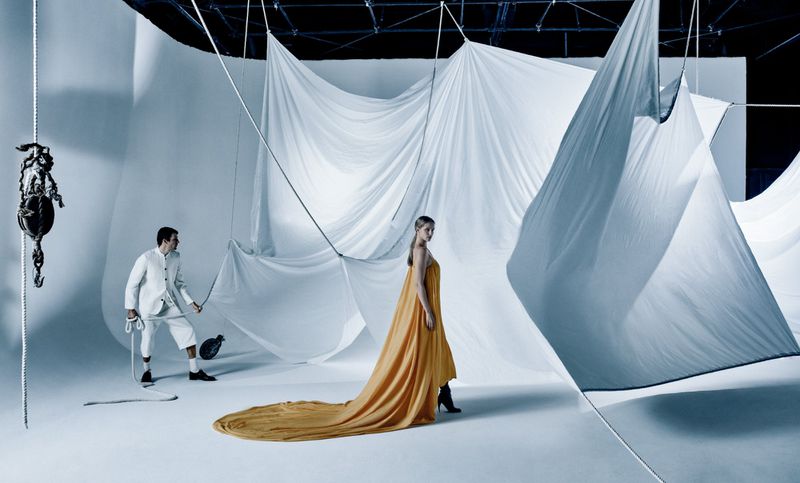 Fashion shot featuring a woman in an orange dress with a long train, accompanied by a man in white pulling on fabric ropes.