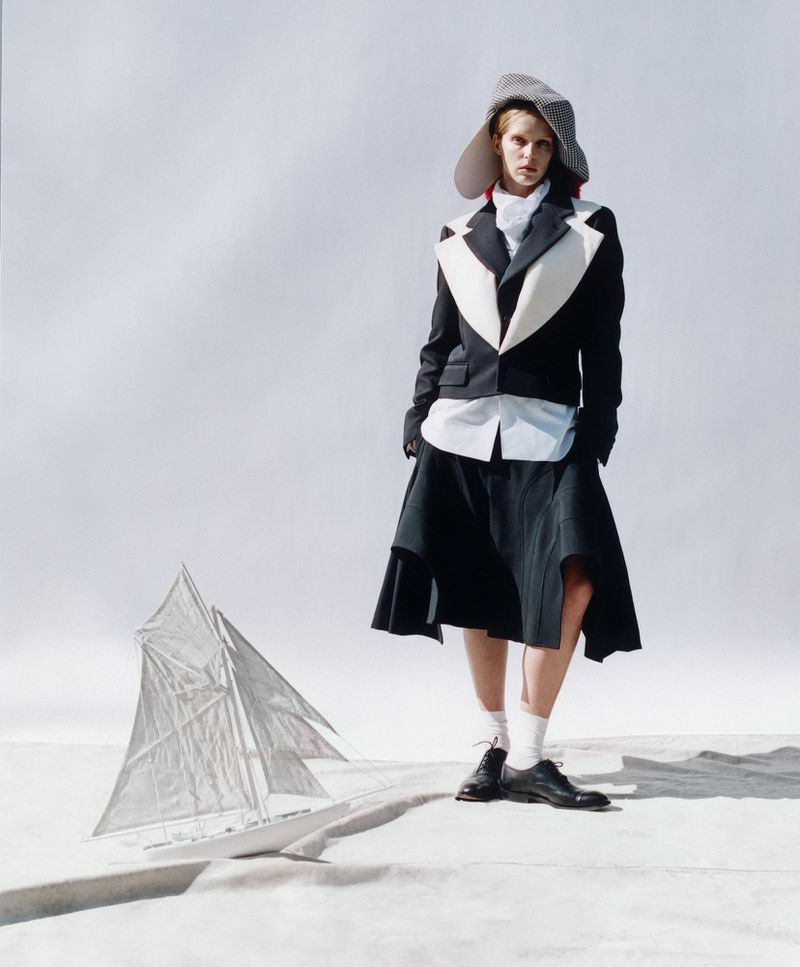 Fashion shot of a person wearing a wide-brim hat, jacket, and skirt, standing near a sailboat on a white background, full shot.