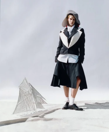 Fashion shot of a person wearing a wide-brim hat, jacket, and skirt, standing near a sailboat on a white background, full shot.