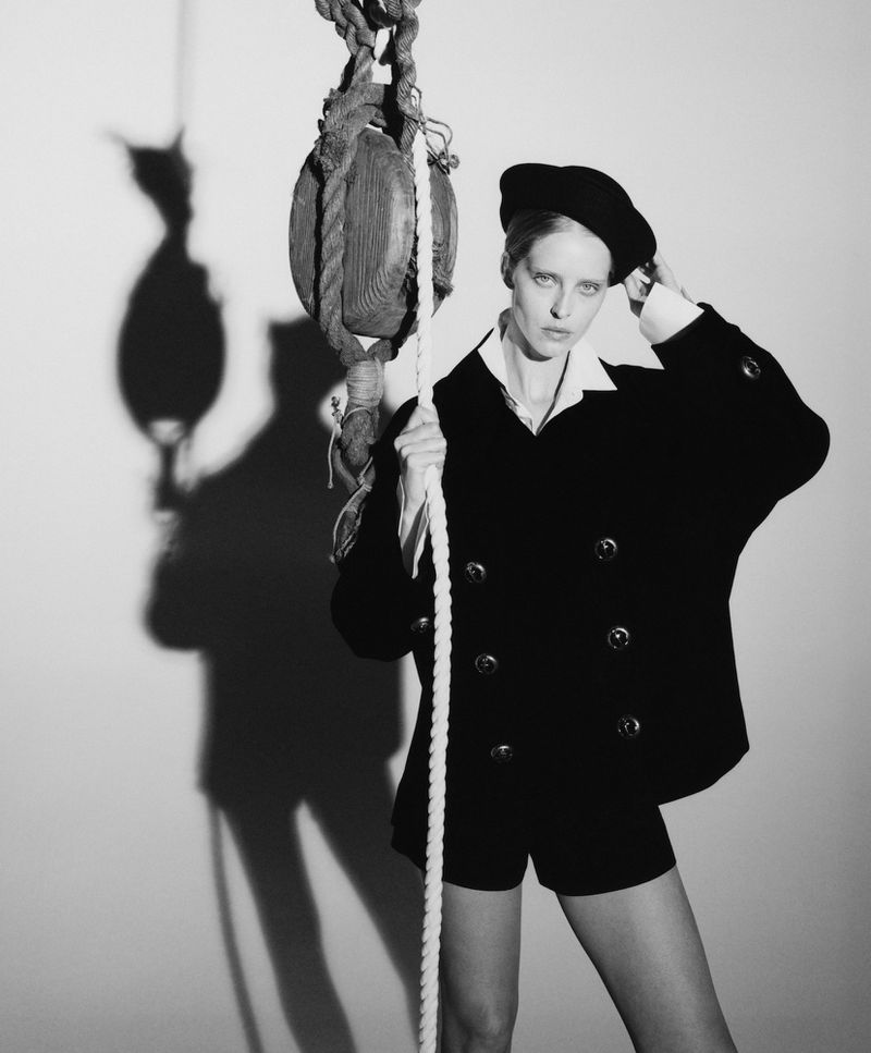 Woman in black jacket, shorts, and beret holding rope next to a wood block and tackle, casting a shadow on the wall.