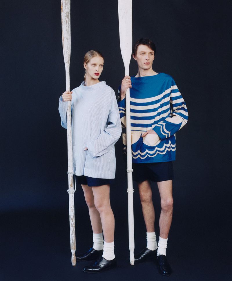 Two models in studio with painted wooden oars, wearing nautical-themed outfits and white socks with shiny black leather shoes against dark backdrop.
