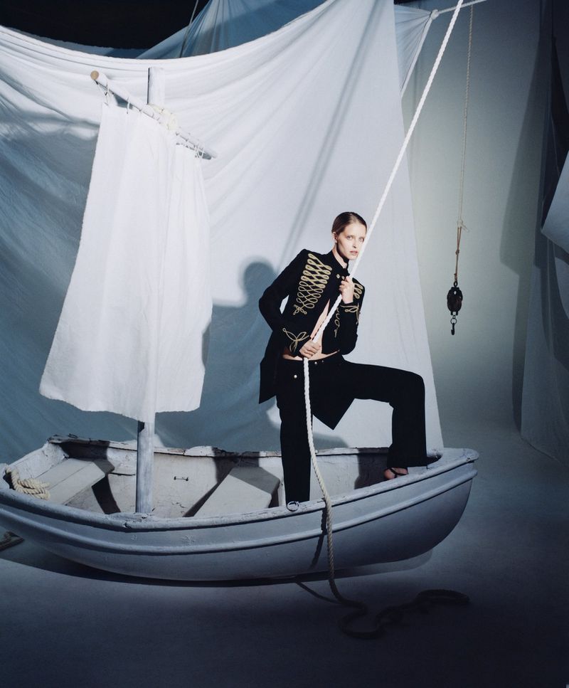 Woman poses in a rowboat with a sail made of white fabric in a studio setting, wearing a black military-style jacket and pants.