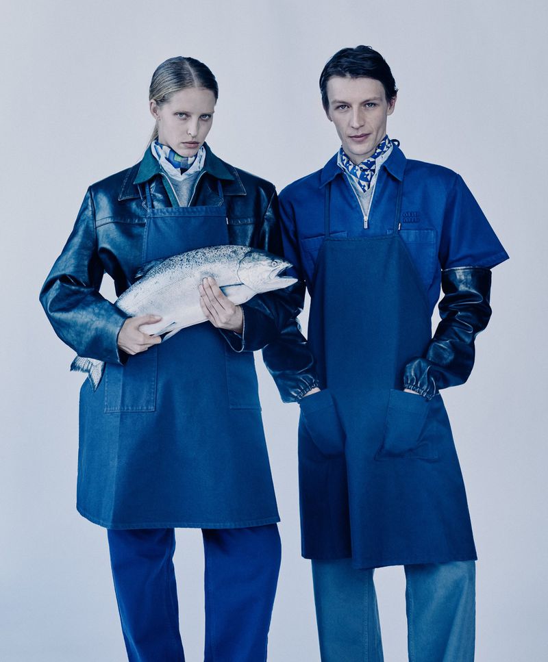 Two models in aprons and gloves pose with a large fresh fish in a studio, captured in a medium shot with neutral lighting.