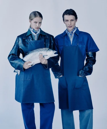Two models in aprons and gloves pose with a large fresh fish in a studio, captured in a medium shot with neutral lighting.