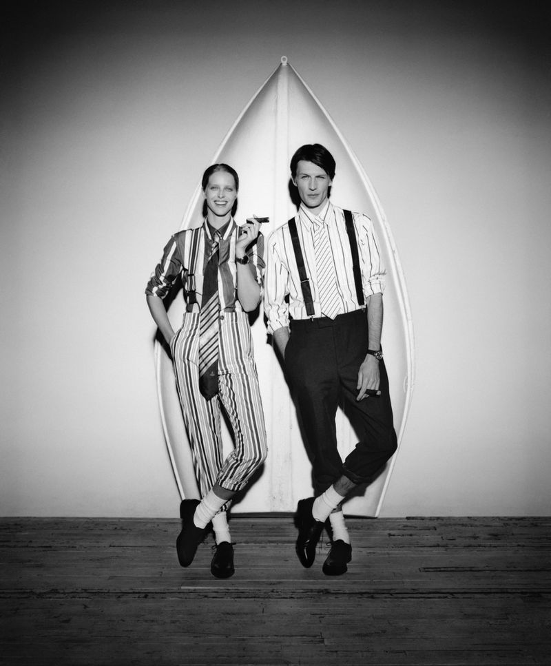 Monochrome image of a stylish couple standing next to a surfboard, woman holding a cigar, both in striped outfits, against a white backdrop.