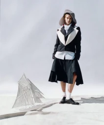Model wearing a black and white tailored jacket, flared skirt, and large brimmed hat standing beside a miniature model sailboat