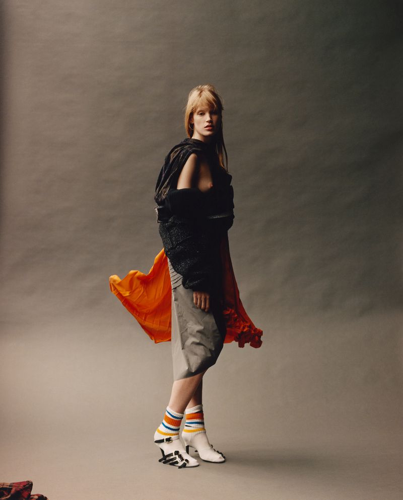 Blonde fashion model in studio poses in a black textured jacket, bright orange skirt, grey shorts, striped socks, and white heels.