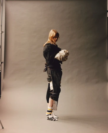 Woman posing in a dark outfit holding white fabric in a photo studio setting with a grey background and studio lighting equipment.