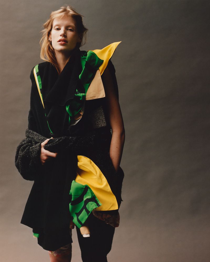 A fair-skinned woman with blonde hair poses in a black outfit with green and yellow accents against a neutral gray backdrop.