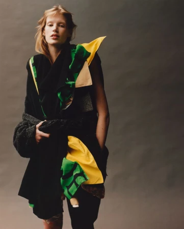 A fair-skinned woman with blonde hair poses in a black outfit with green and yellow accents against a neutral gray backdrop.