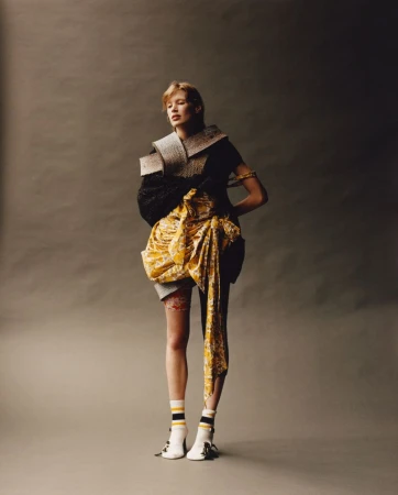 Model standing against a grey seamless backdrop wearing avant-garde textured clothing with a yellow wrap skirt and striped socks