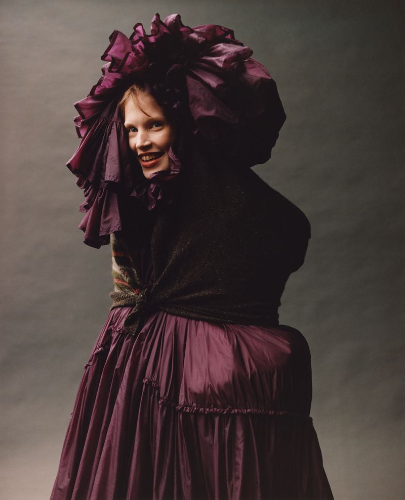 Woman in a large purple ruffled hat and dress smiles in a studio portrait against a grey backdrop, fashion and costume forward.
