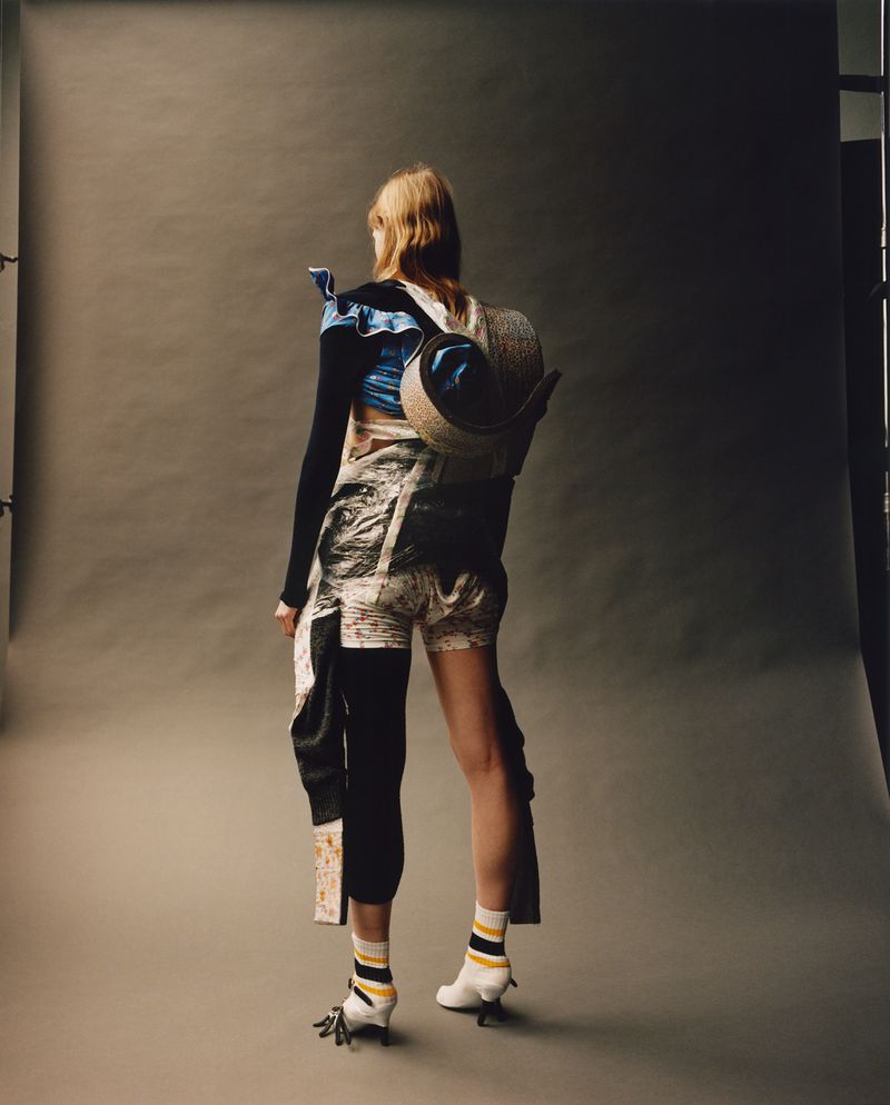Woman stands wearing avant-garde fashion with straw hat backpack in a studio setting, captured in a back view portrait.