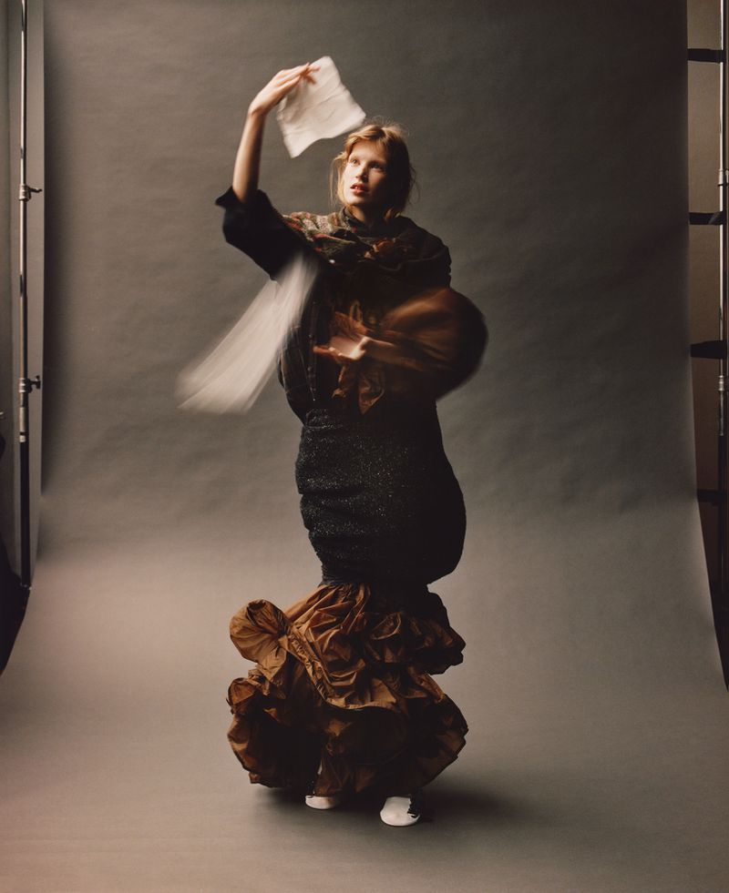 Full-length shot of a female fashion model in an ornate black and brown gown holding a white paper in a studio setting.