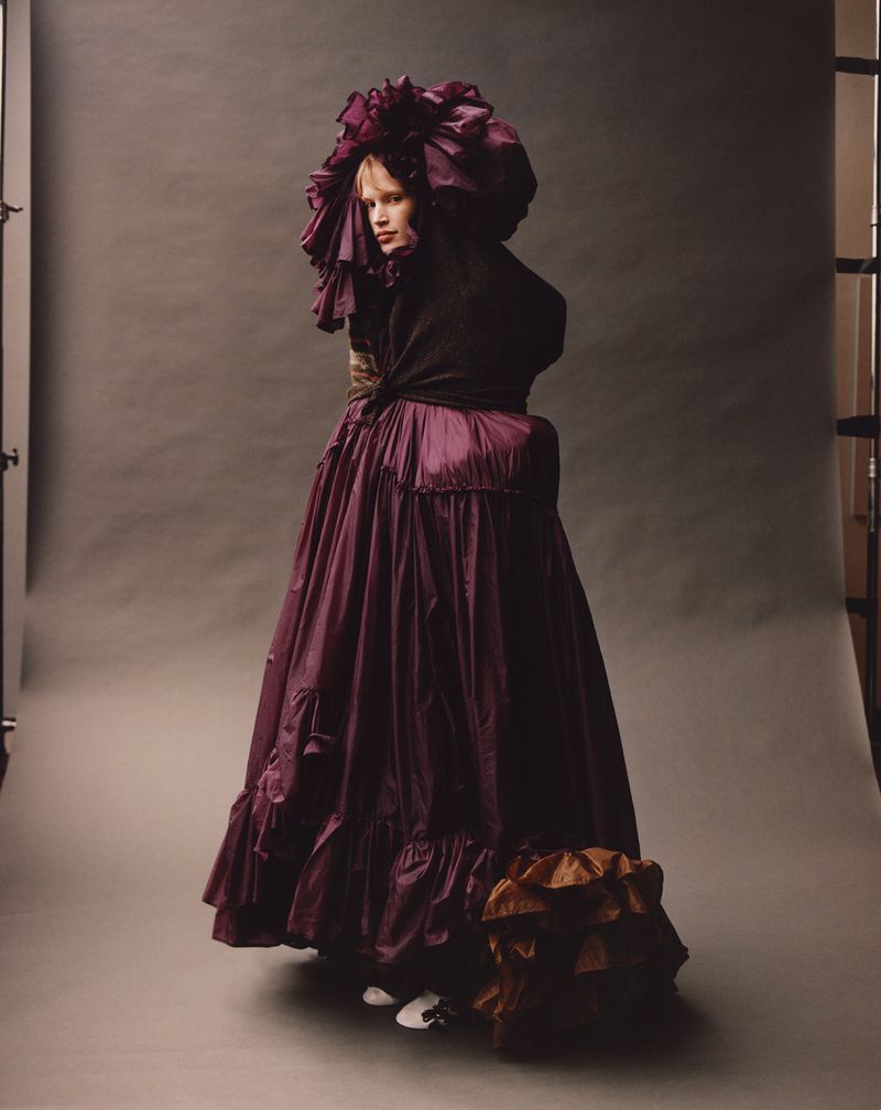 A fashion portrait of a woman in a large purple dress and headpiece, posing in a studio setting with soft lighting and a neutral background.