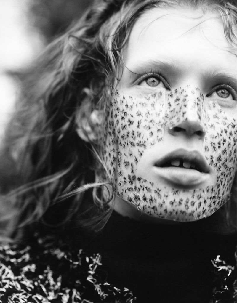 Close-up black and white portrait of a young woman with freckles, curly hair, looking upward with a blurred background, wearing a dark top.