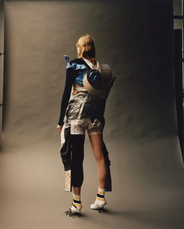 Model standing facing away wearing avant-garde patterned clothing and heels in a studio with grey seamless backdrop.