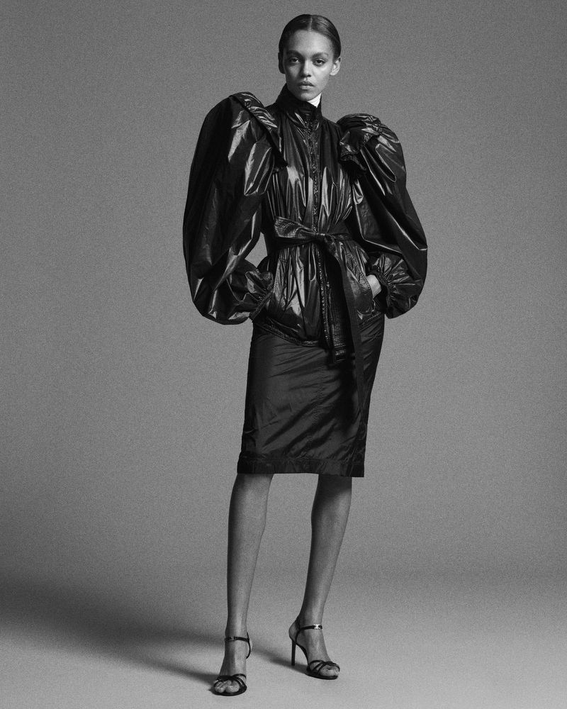Model Bebe Parnell wearing a shiny belted black puff-sleeve dress against a grey studio backdrop.
