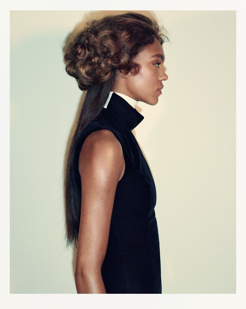 Profile shot of a Black woman with styled brown hair wearing a sleeveless black top with a white turtleneck against a light yellow background.