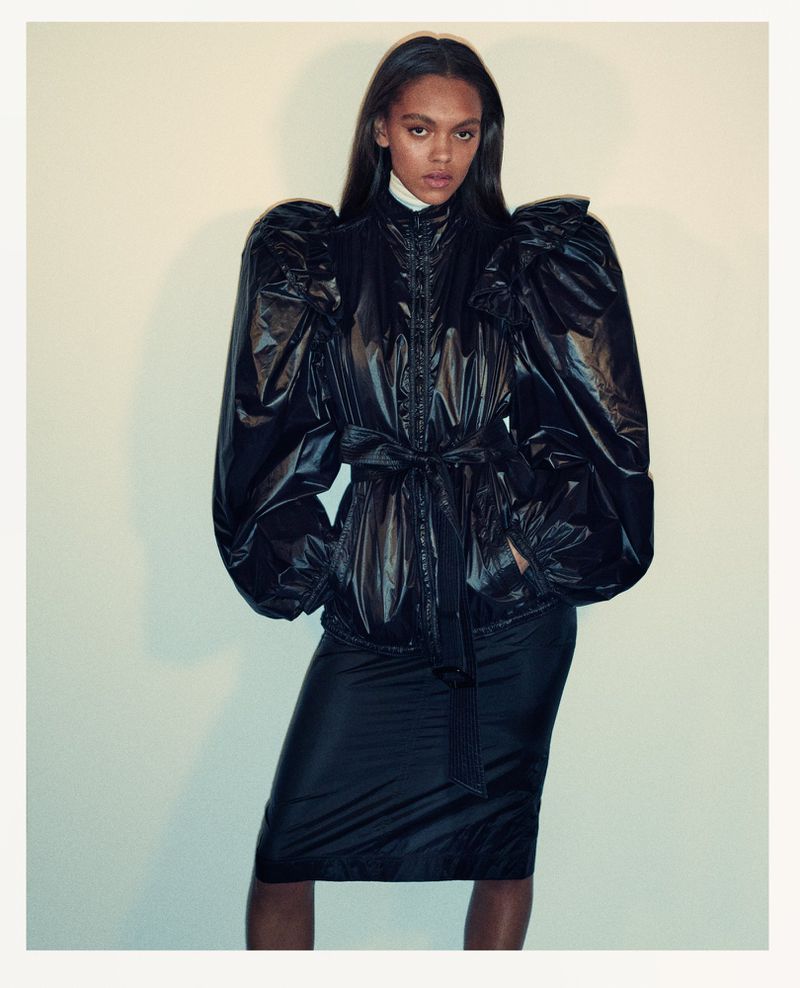 Fashionable woman in a black puffer coat and skirt posing in front of a neutral background. Fashion editorial photography, studio shot.