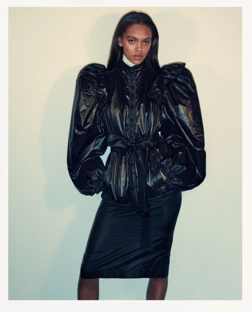 Fashionable woman in a black puffer coat and skirt posing in front of a neutral background. Fashion editorial photography, studio shot.