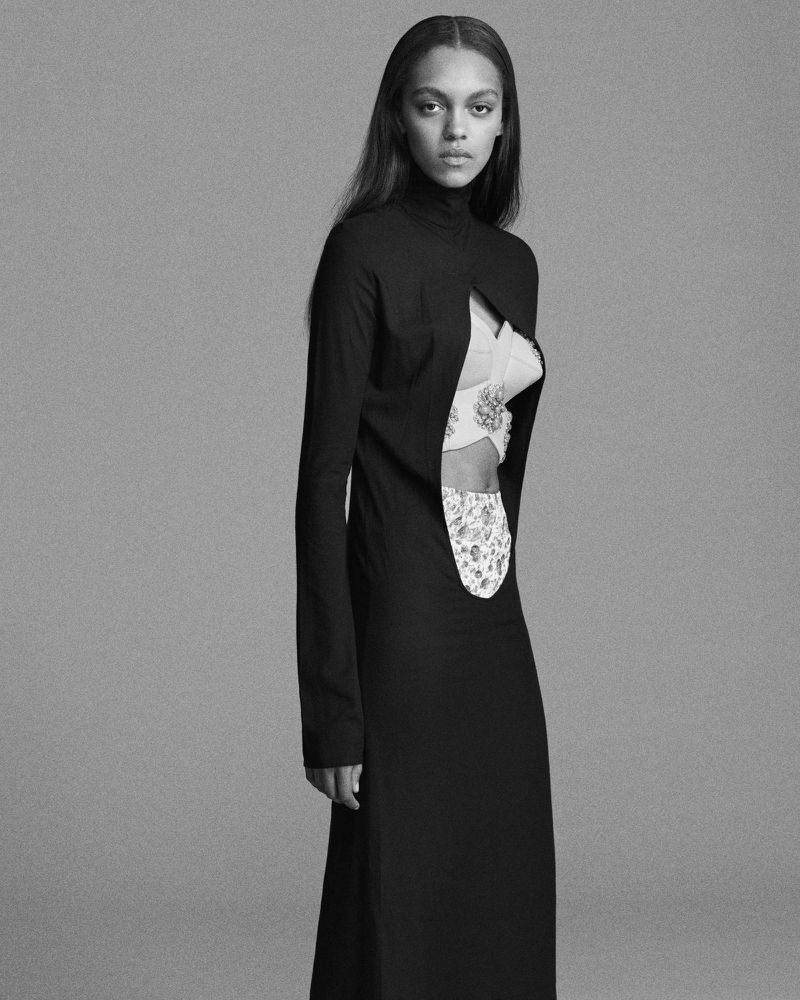 Elegant fashion shot of an adult female model posing in a black high-neck dress with floral print details against a grey backdrop.