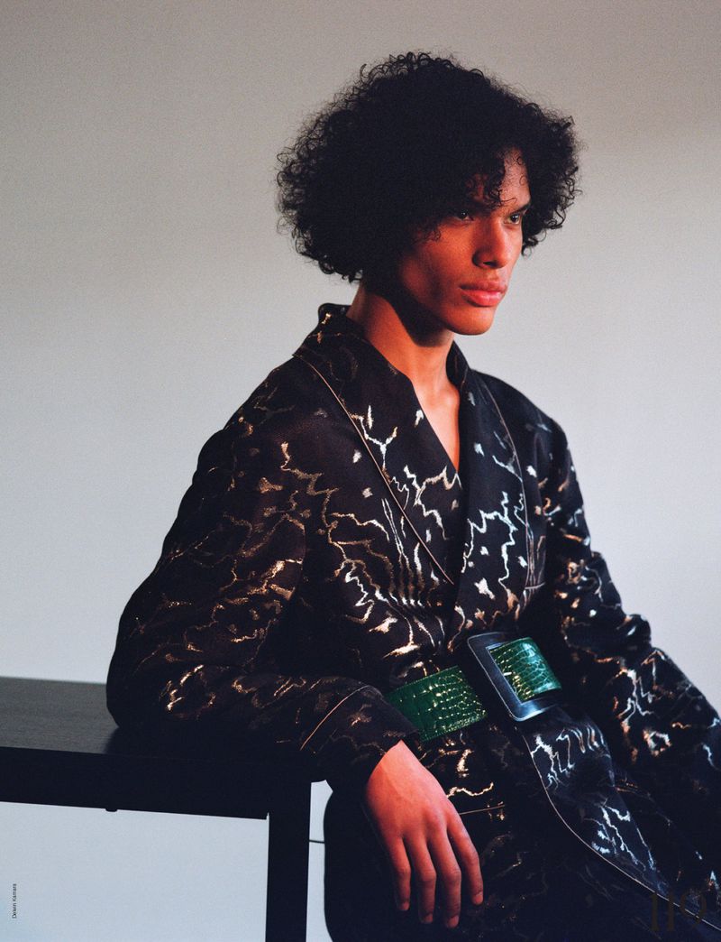 An adult male with dark curly hair poses indoors wearing a robe with a metallic pattern and a green belt, leaning on a dark table.