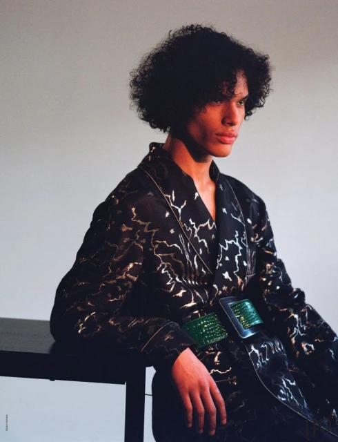 An adult male with dark curly hair poses indoors wearing a robe with a metallic pattern and a green belt, leaning on a dark table.