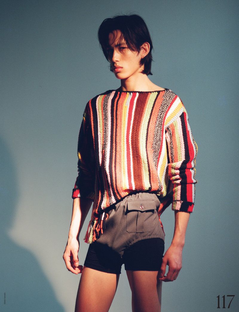 An Asian male model poses in a studio wearing a colourful striped sweater and shorts against a light-blue backdrop, showcasing high fashion.