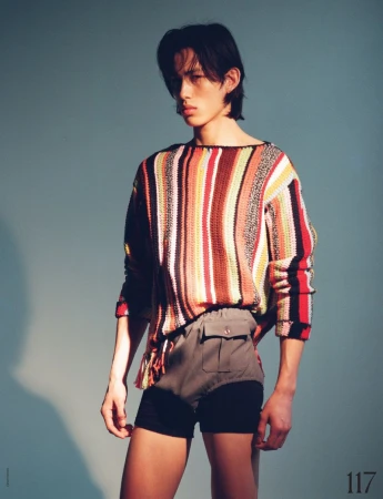 An Asian male model poses in a studio wearing a colourful striped sweater and shorts against a light-blue backdrop, showcasing high fashion.