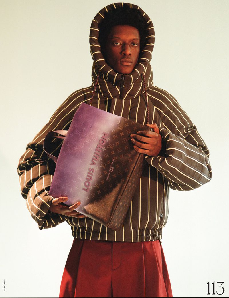 Fashion shot of a Black male model in a striped puffer jacket holding a Louis Vuitton bag against a neutral studio backdrop.
