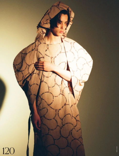 A model poses in a hooded tan patterned dress in a studio with directional lighting, fashion photography, style, and artistic composition.