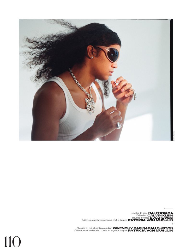 A person with long dark hair is captured in a studio shot, wearing sunglasses and a white tank top, accessorized with a silver chain.