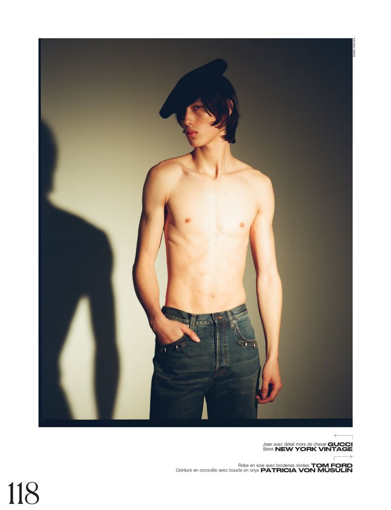 Fashion portrait of a shirtless male model wearing jeans and a beret, posing against a neutral-toned background with a visible shadow.