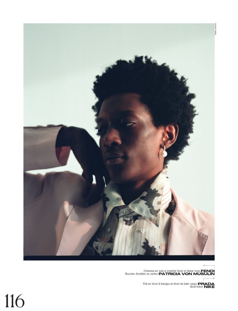 Portrait of a Black male model in a pink blazer and patterned shirt, wearing a pearl earring, posing indoors against a white background.