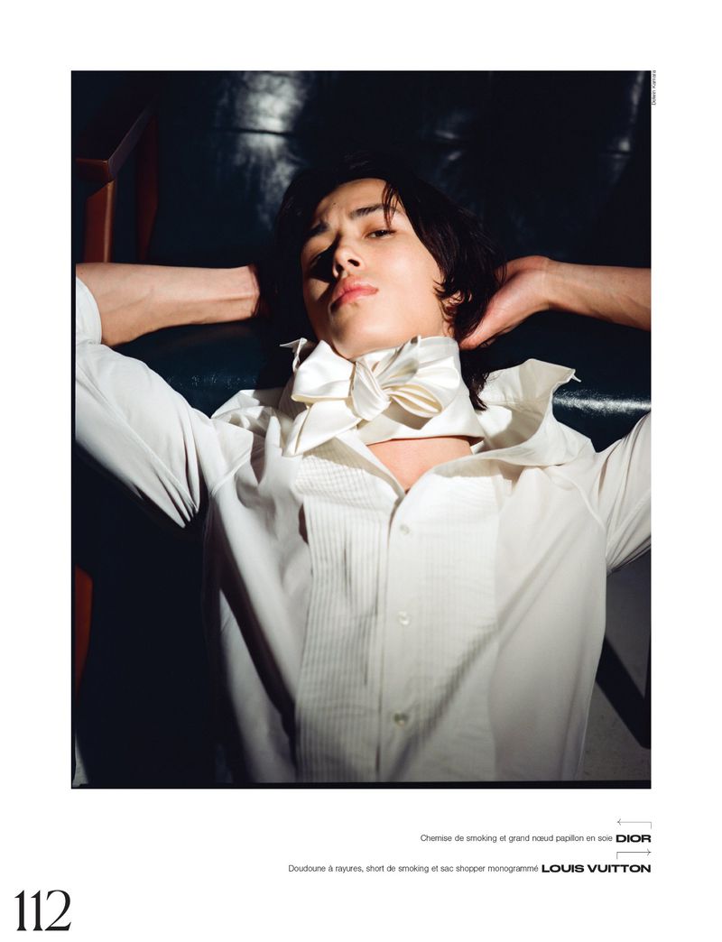 A single, adult male model with dark hair reclines in a white shirt and large bow tie, arms above his head, in a dimly lit, indoor setting.