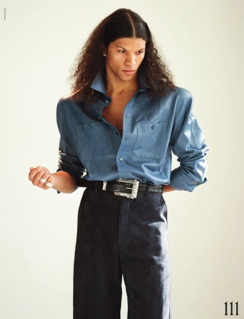 A stylish young adult man with long, wavy brown hair wears a blue button-down shirt and dark trousers in a brightly lit studio portrait.